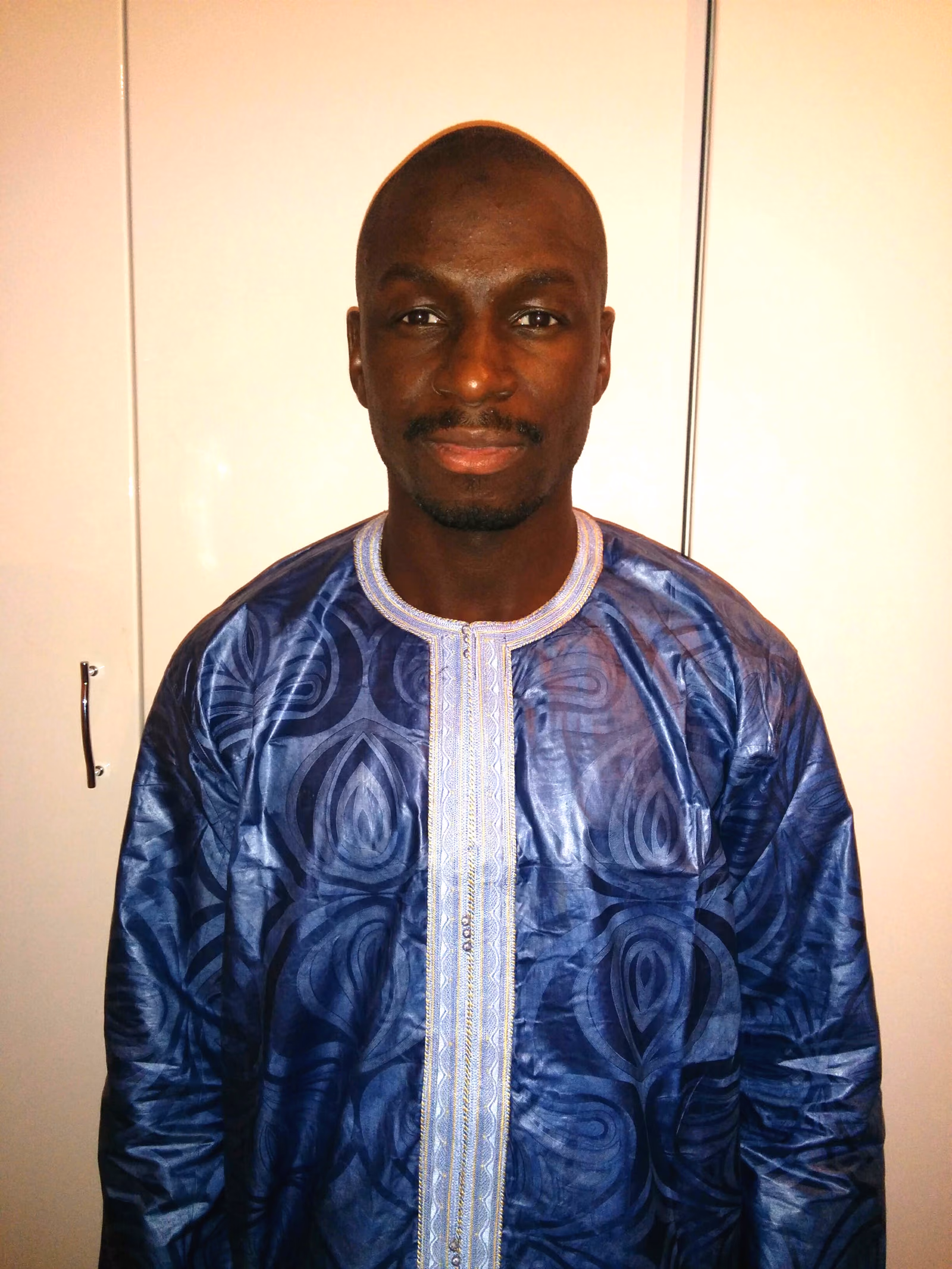 A man in a deep blue bazin boubou, pale embroidery running down the chest, the wide sleeves falling to either side.