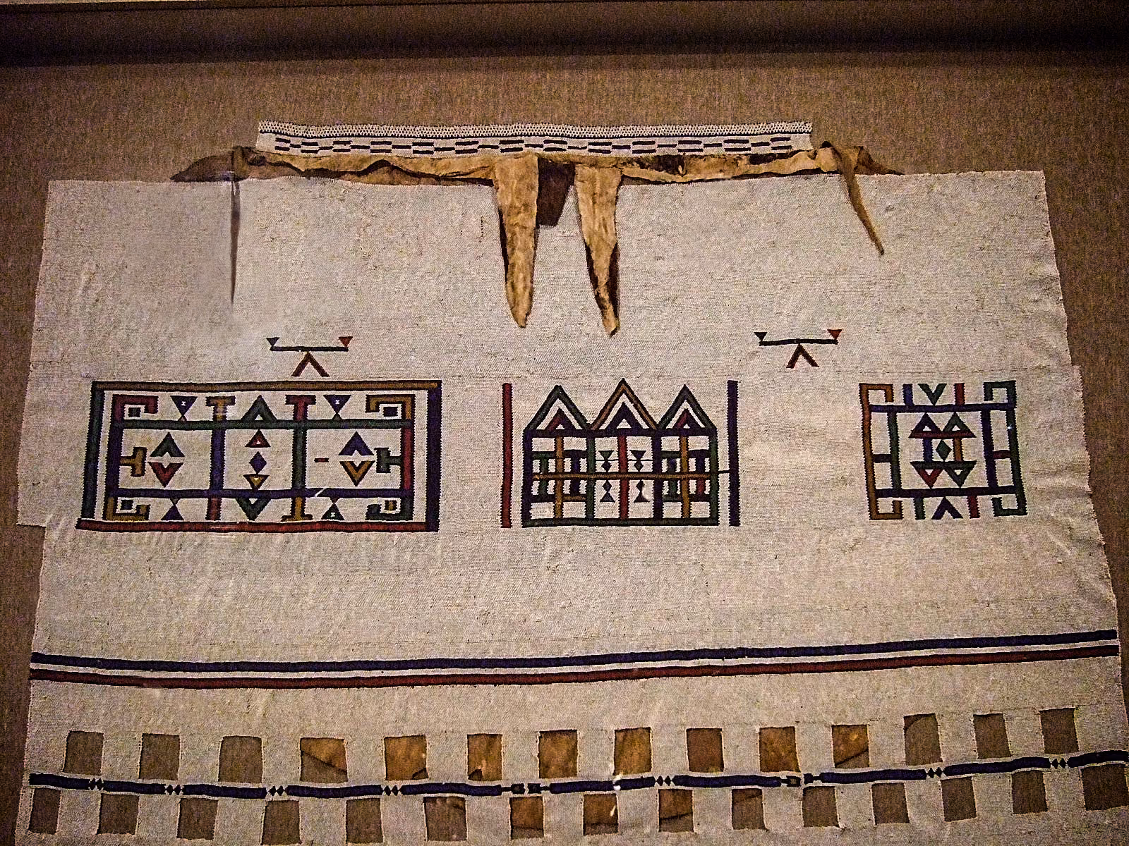 A Ndebele linaga: a beaded cape on a pale ground, worked with three panels of dark blue, red and gold geometric figures above a row of beaded fringe.