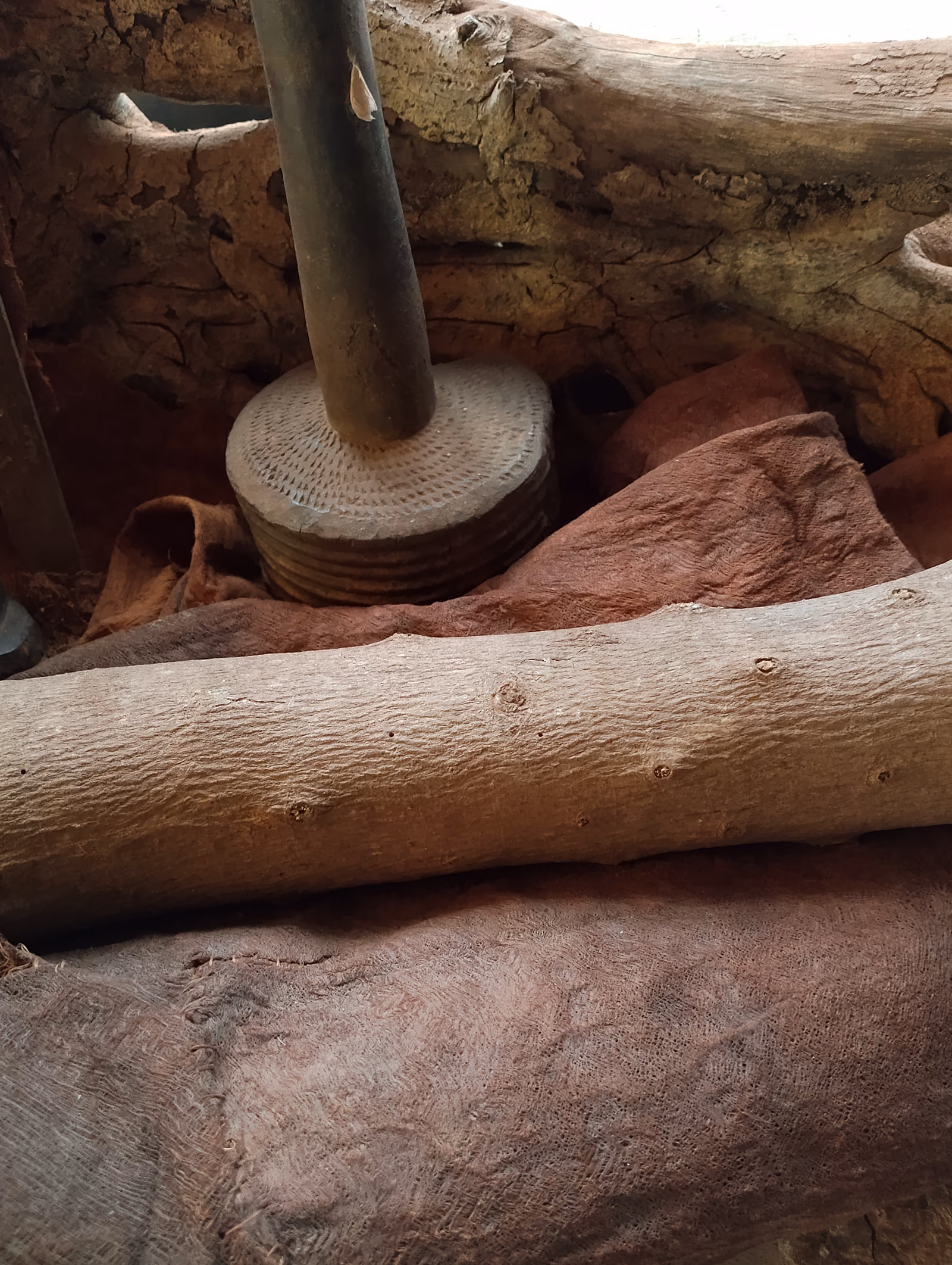 A grooved wooden mallet resting on a cut log beside a folded sheet of russet bark cloth.