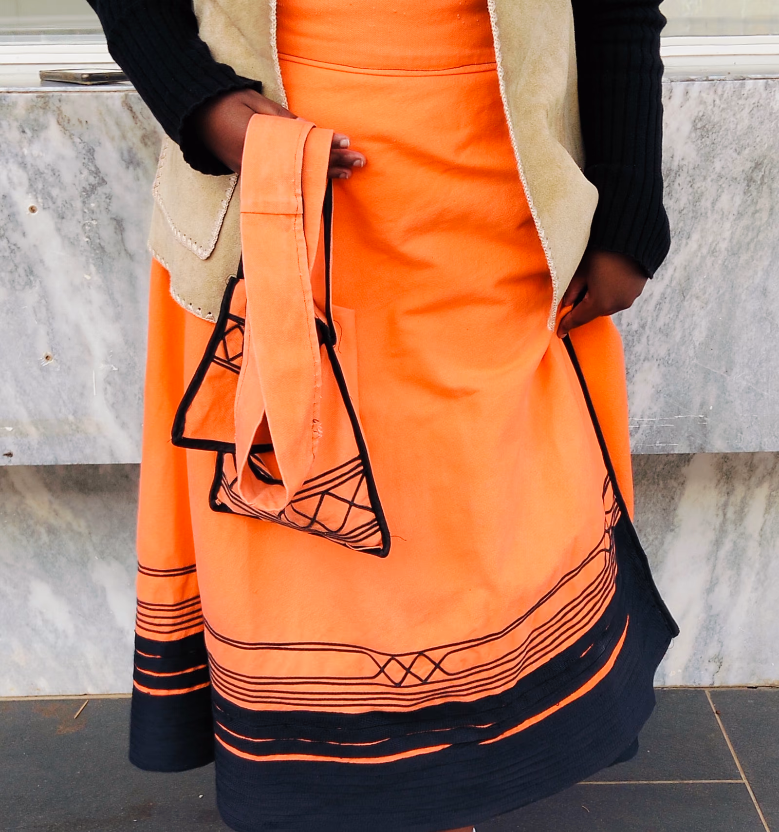 An orange umbhaco garment trimmed with bands of black braid, displayed on a stand.