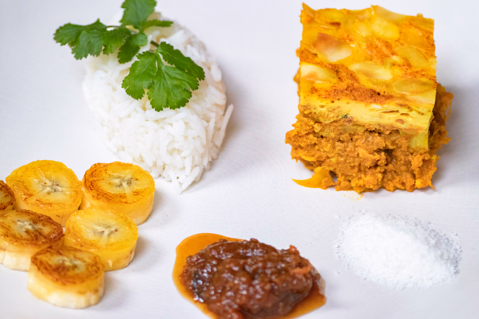 A square of baked bobotie with a set golden egg topping over spiced mince, plated with white rice, fried plantain, chutney and desiccated coconut.