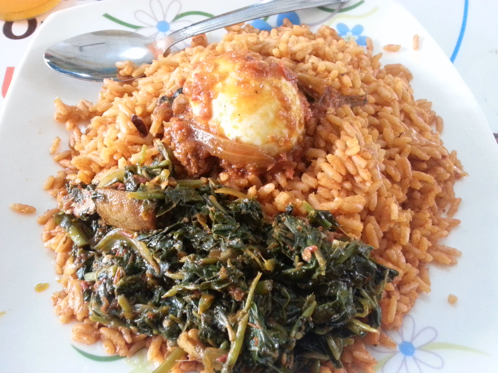 A plate of deep orange-red jollof rice served with cooked leafy greens and a piece of chicken topped with a boiled egg.