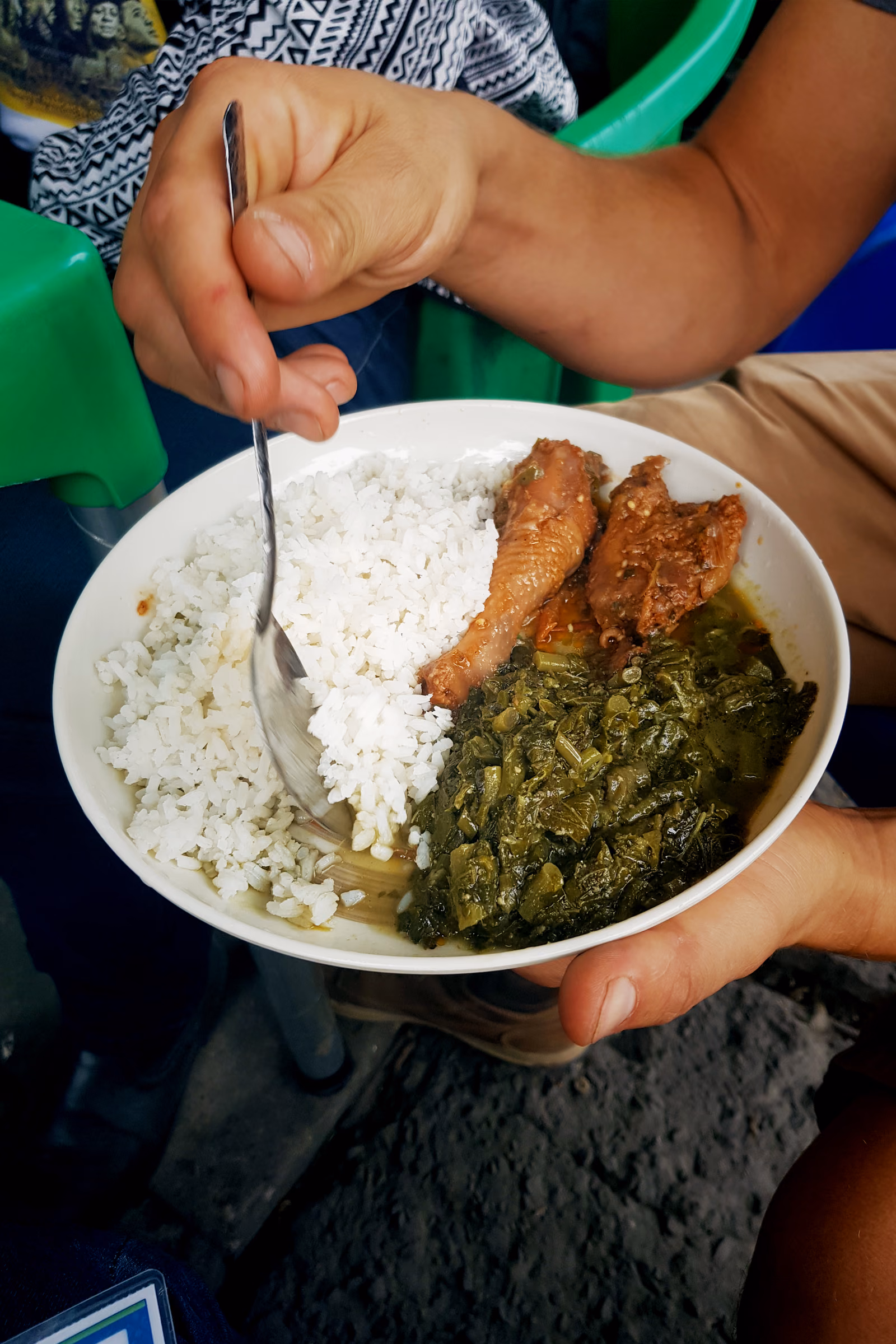 A plate of white rice and dark stewed greens beside chicken in a thick reddish palm-nut sauce.