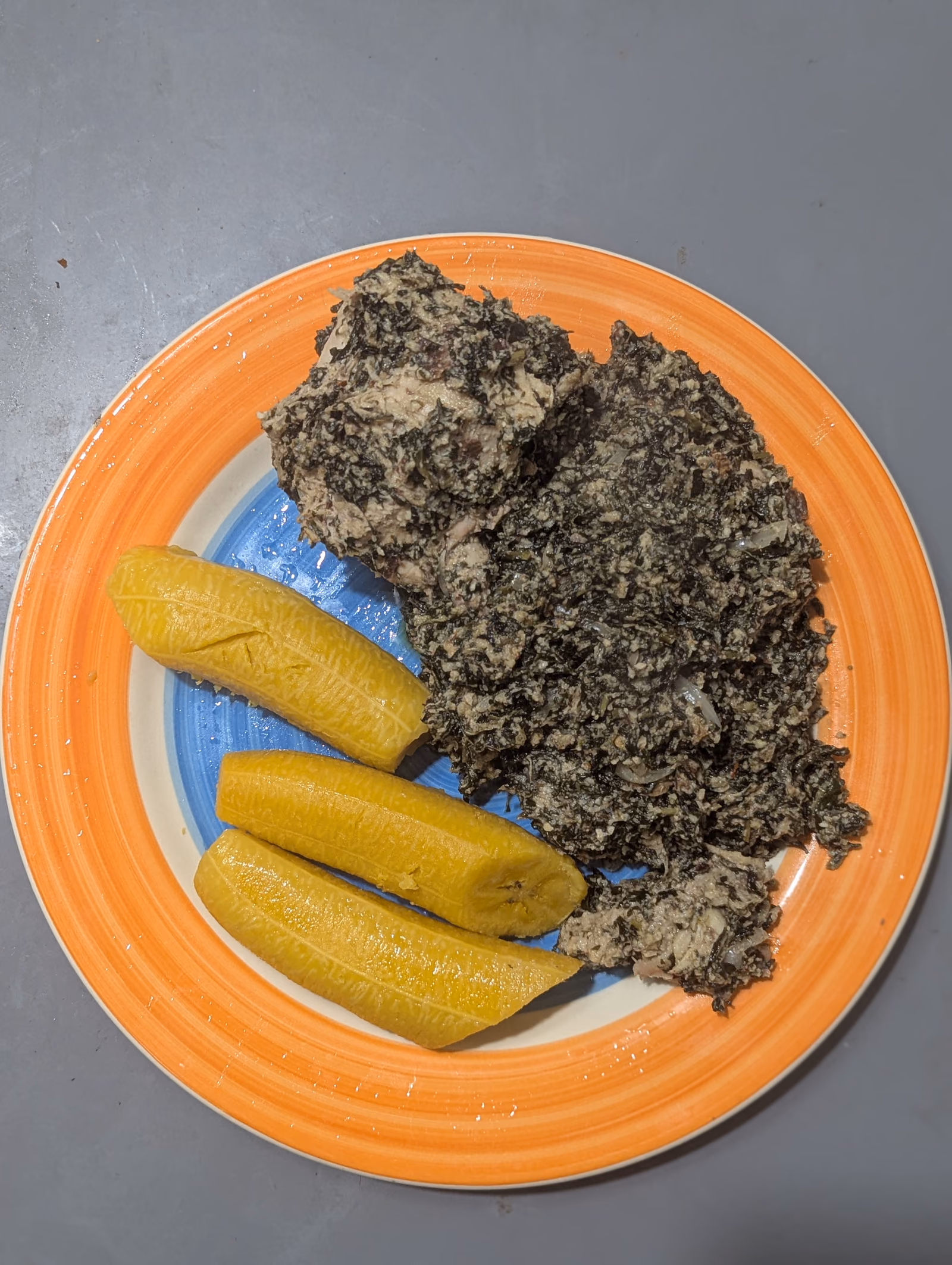 A plate of ndole, finely chopped dark bitterleaf greens cooked in a pale groundnut sauce, served beside two lengths of boiled ripe plantain.